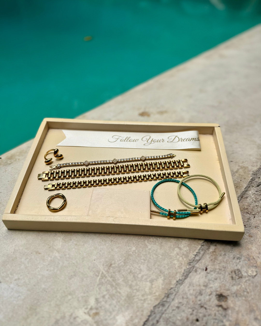 Tray of luxury jewelry displayed poolside featuring the Modern Muse Ring, gold Rolex-style link bracelets, a clover tennis bracelet, and colorful H cord adjustable rope bracelets.