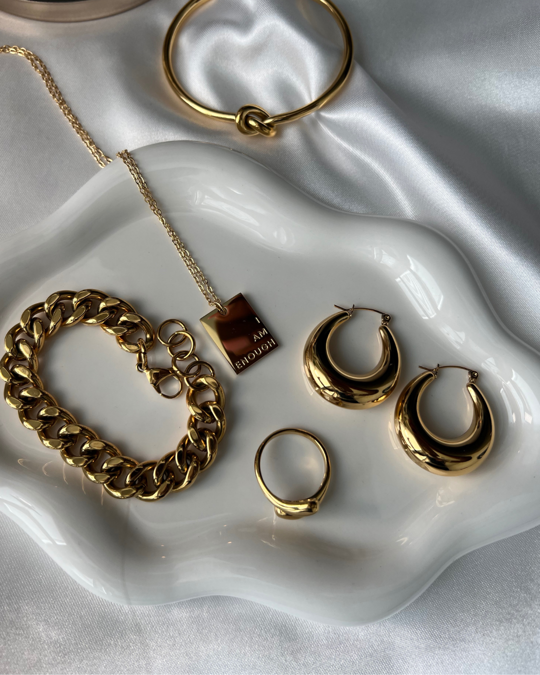 Flat lay of gold jewelry including the I Am Enough necklace, chunky bracelet, hoop earrings, and minimalist ring display on a wavy ceramic tray with satin background.