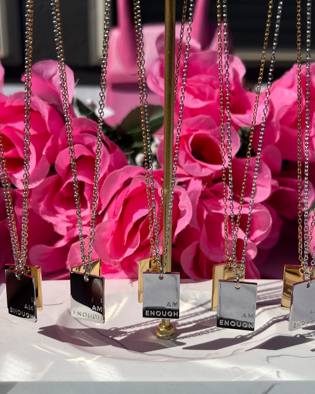 Multiple I Am Enough necklaces in gold and silver shine boldly against pink roses, styled to affirm self-worth and remind women everywhere that they are more than enough--every single day.