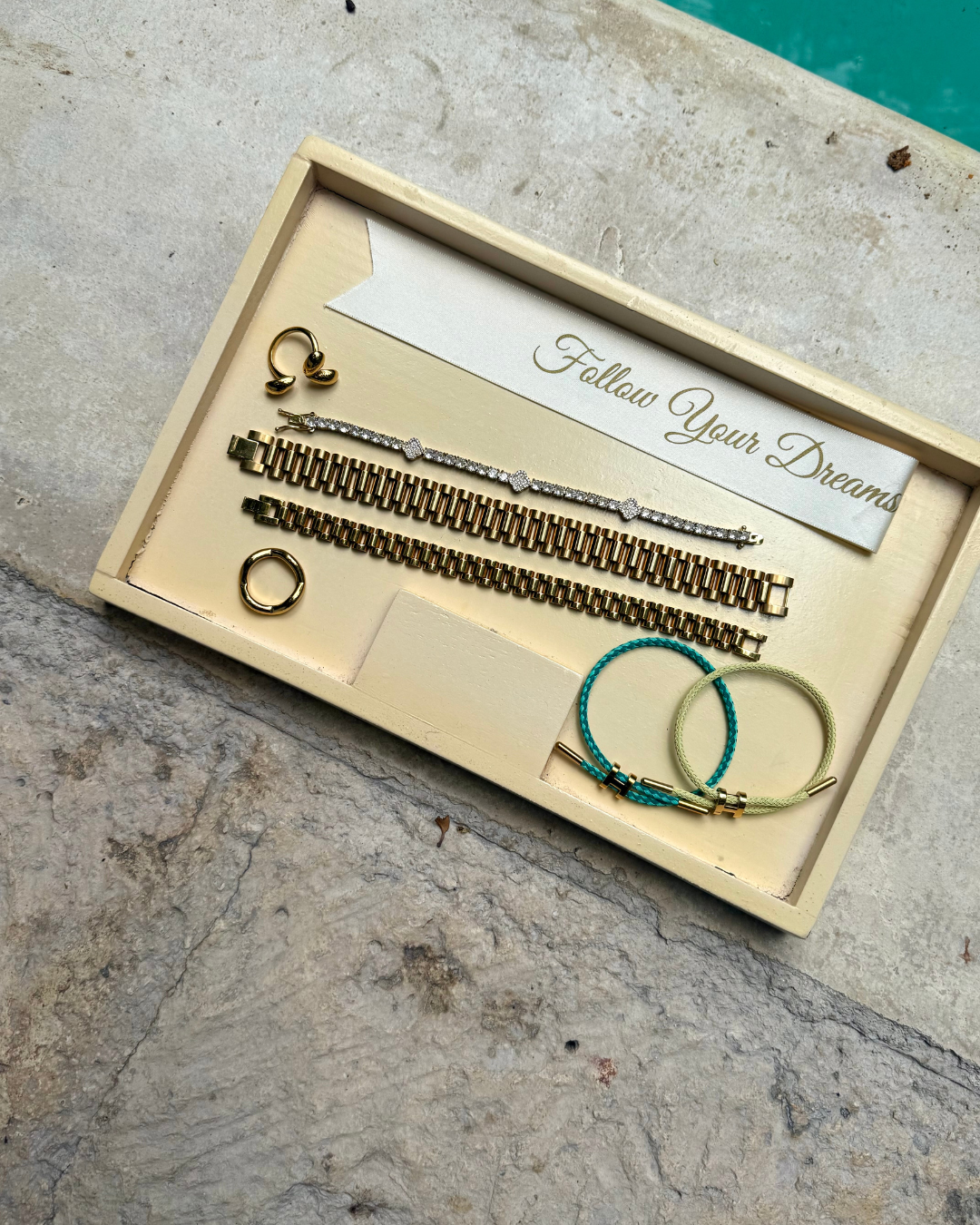 Flat lay of gold and colorful bracelets, rings, and earrings displayed on a tray with a “Follow Your Dreams” card beside a pool — perfect summer travel jewelry inspiration.
