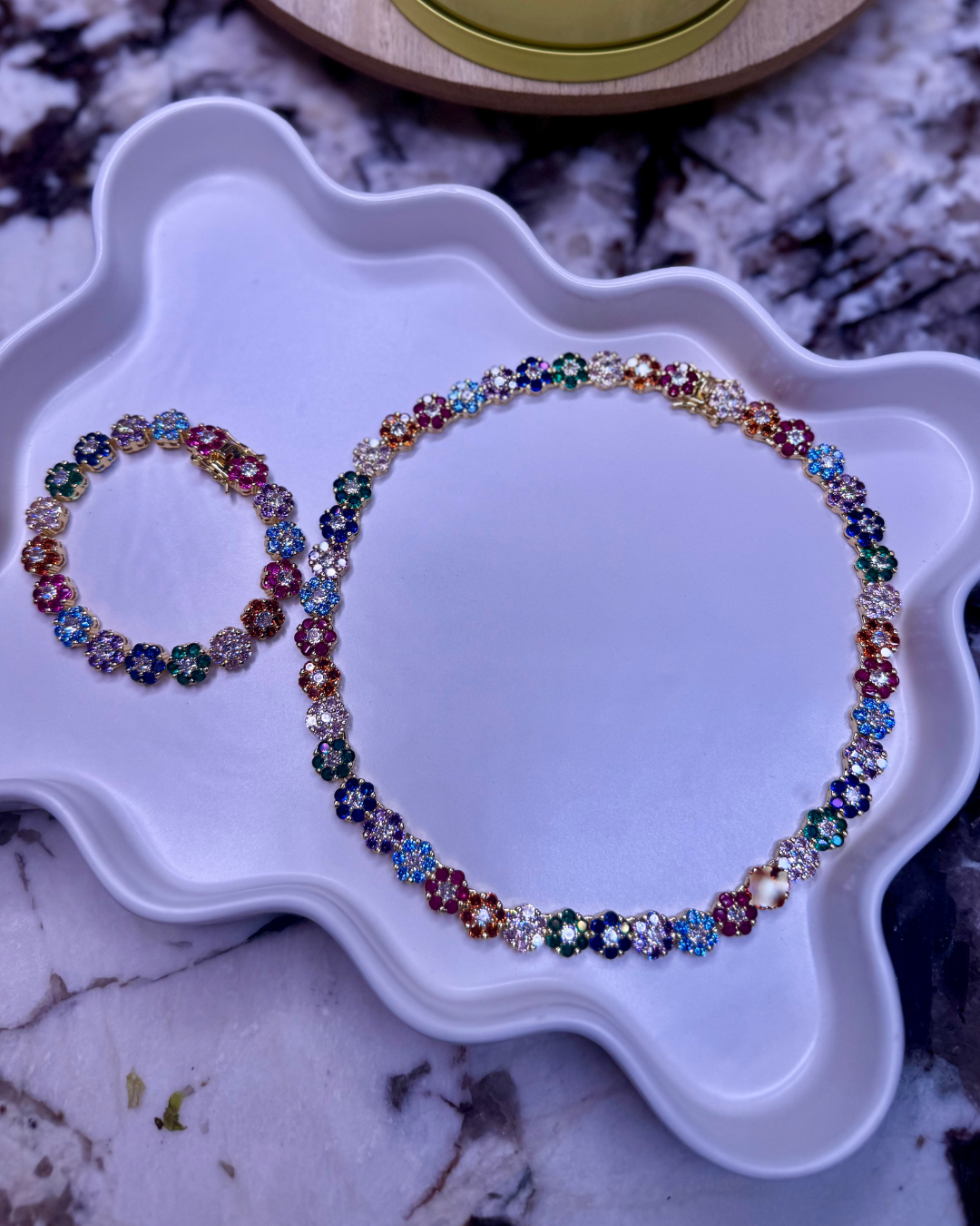 In Full Bloom Bracelet – multicolor CZ floral cluster bracelet in 18k gold and silver stainless steel, displayed on a white tray with the matching necklace and a marble background.