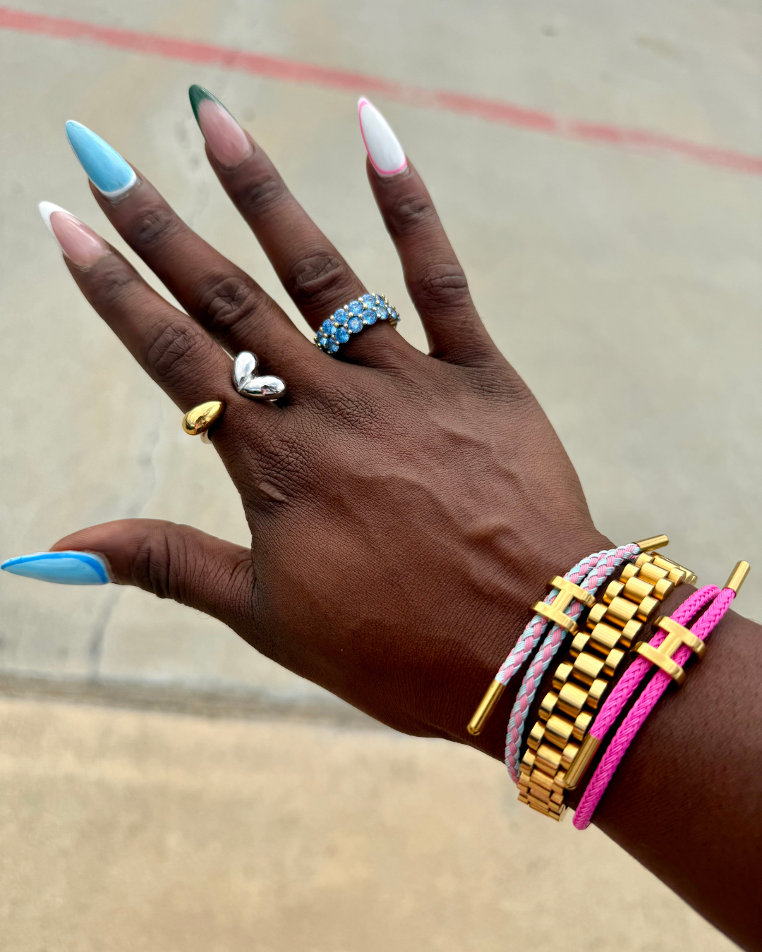 Close-up of a hand with fun, colorful nails wearing the Modern Muse Ring in a gold and silver tear-drop design, styled with Rolex-style bracelets and H cord adjustable rope bracelets.