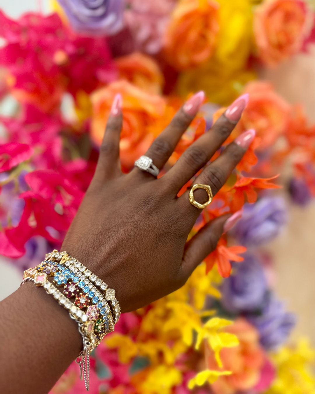 Close-up of a hand wearing the Opulence 18k gold-plated oval-shaped ring, paired with stacked multicolor tennis bracelets, against a vibrant floral background of orange, pink, and purple blooms.