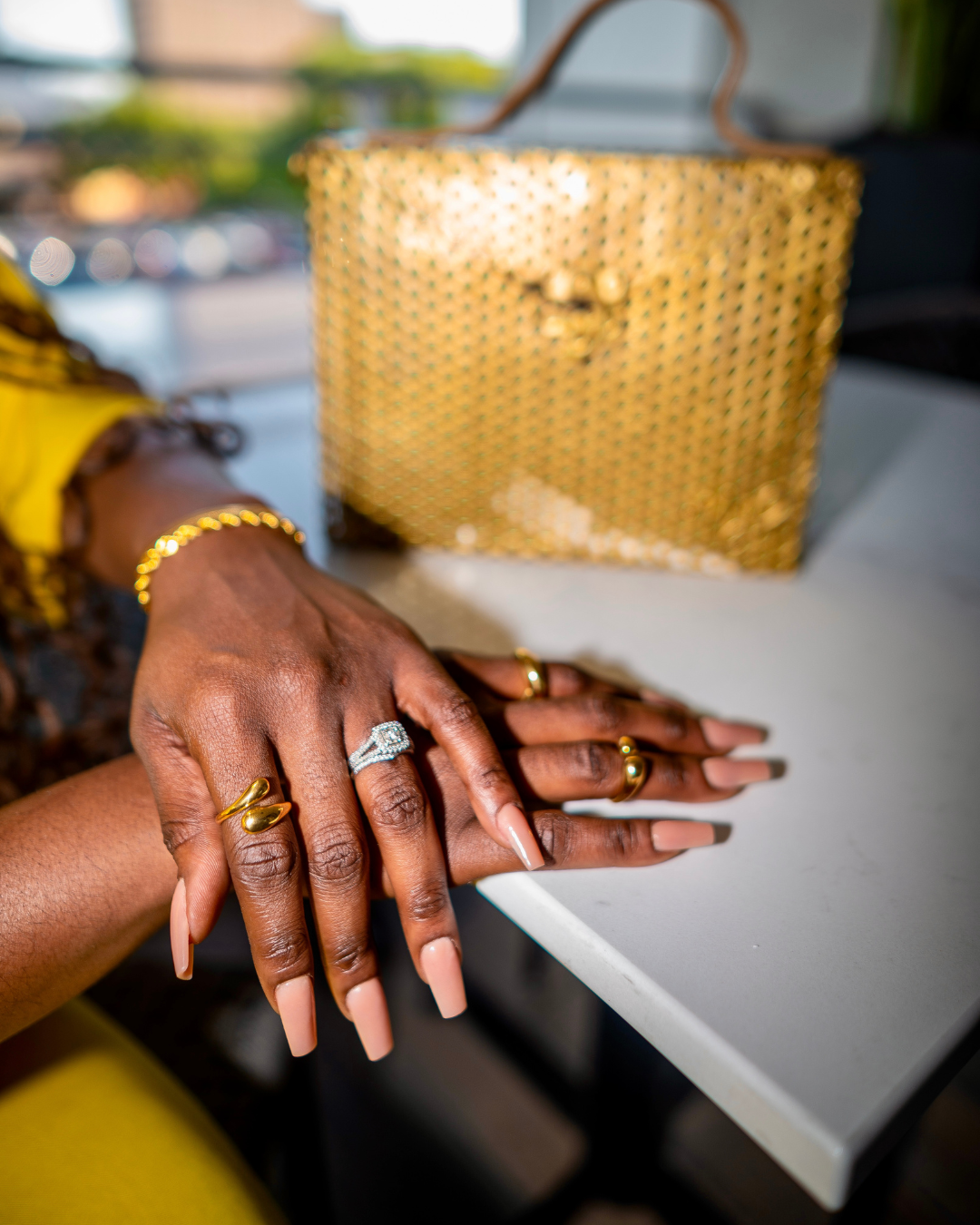 Close-up of woman's hands styled with multiple gold rings, including the Rich AF sculptural ring, against a modern white table with a woven gold handbag in the background. 