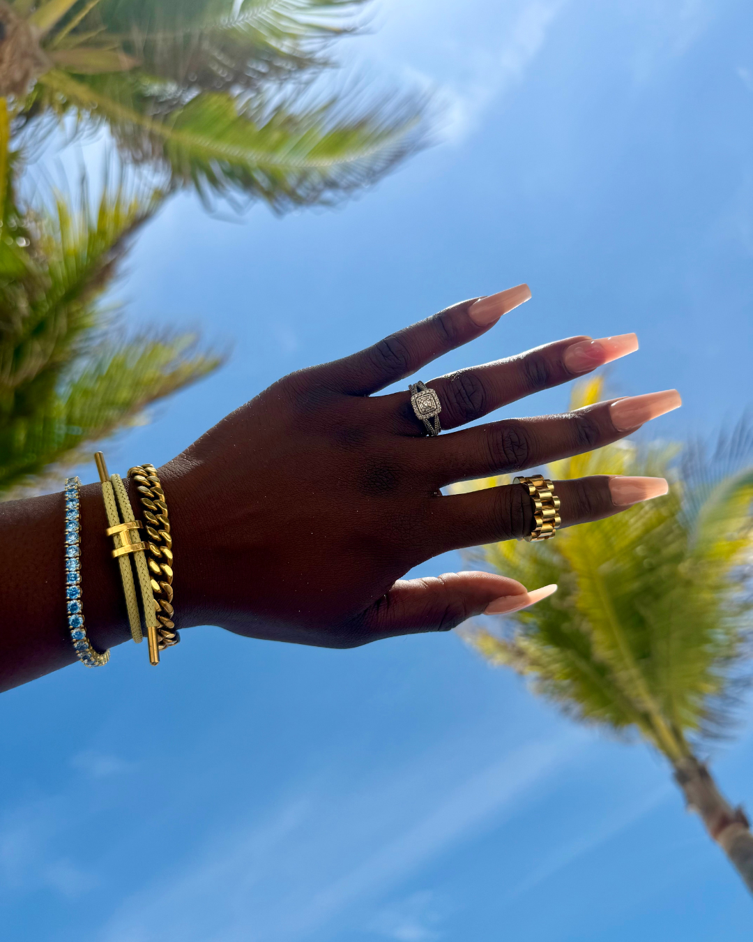 Close-up of a hand against a blue sky and palm trees wearing the Stacked Like a Horse Bracelet—gold bracelet with H-shaped symbol—paired with the Luxe Link Bracelet and Sneaky Link Ring.
