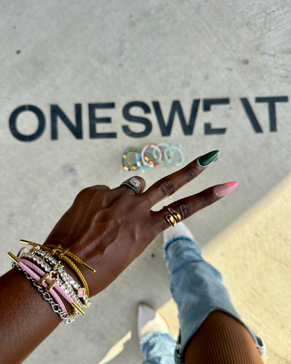 Close-up of a hand flashing a peace sign while wearing stacked Like a Horse Bracelets, the Rich AF Ring, and Lucky Charm Bracelets, styled with pink and green nails, against a concrete floor with “ONESWEAT” in bold black text.