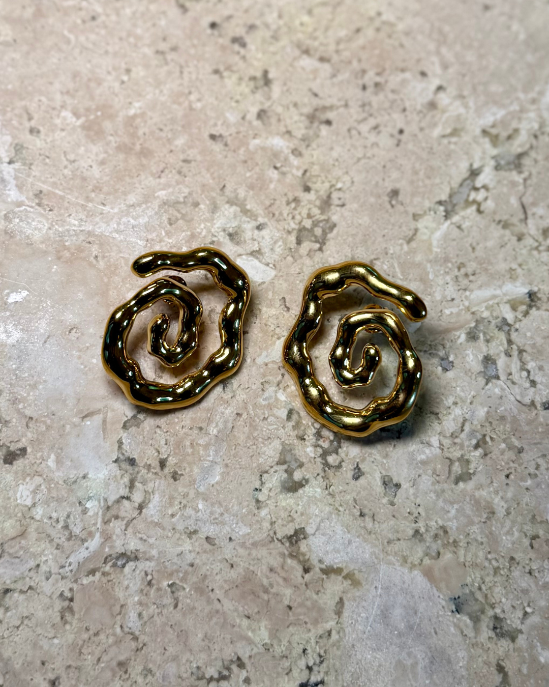 Flat lay of the Twist of Faith Earrings in gold, featuring bold abstract spiral shapes with a glossy finish, displayed on a textured stone surface.