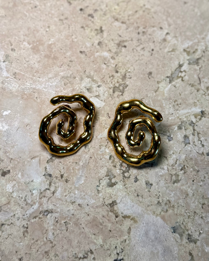 Flat lay of the Twist of Faith Earrings in gold, featuring bold abstract spiral shapes with a glossy finish, displayed on a textured stone surface.