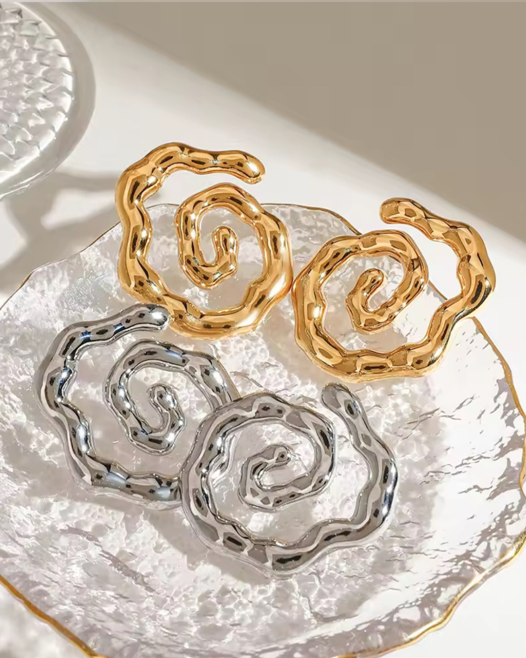 Flat lay of the Twist of Faith Earrings in gold and silver, featuring bold abstract spiral designs displayed on a textured glass dish.