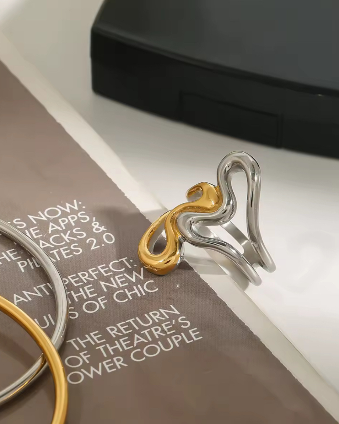 Editorial photo of the Untamed Ring in intertwined gold and silver stainless steel, styled on a magazine with bangles nearby—bold, modern statement jewelry.