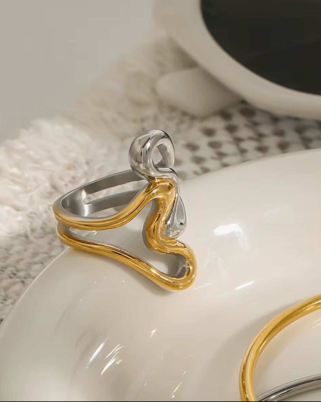 The Untamed Ring in intertwined gold and silver mixed metals, displayed on a white surface, showcasing its sculptural, fluid design that embodies bold elegance and modern style.