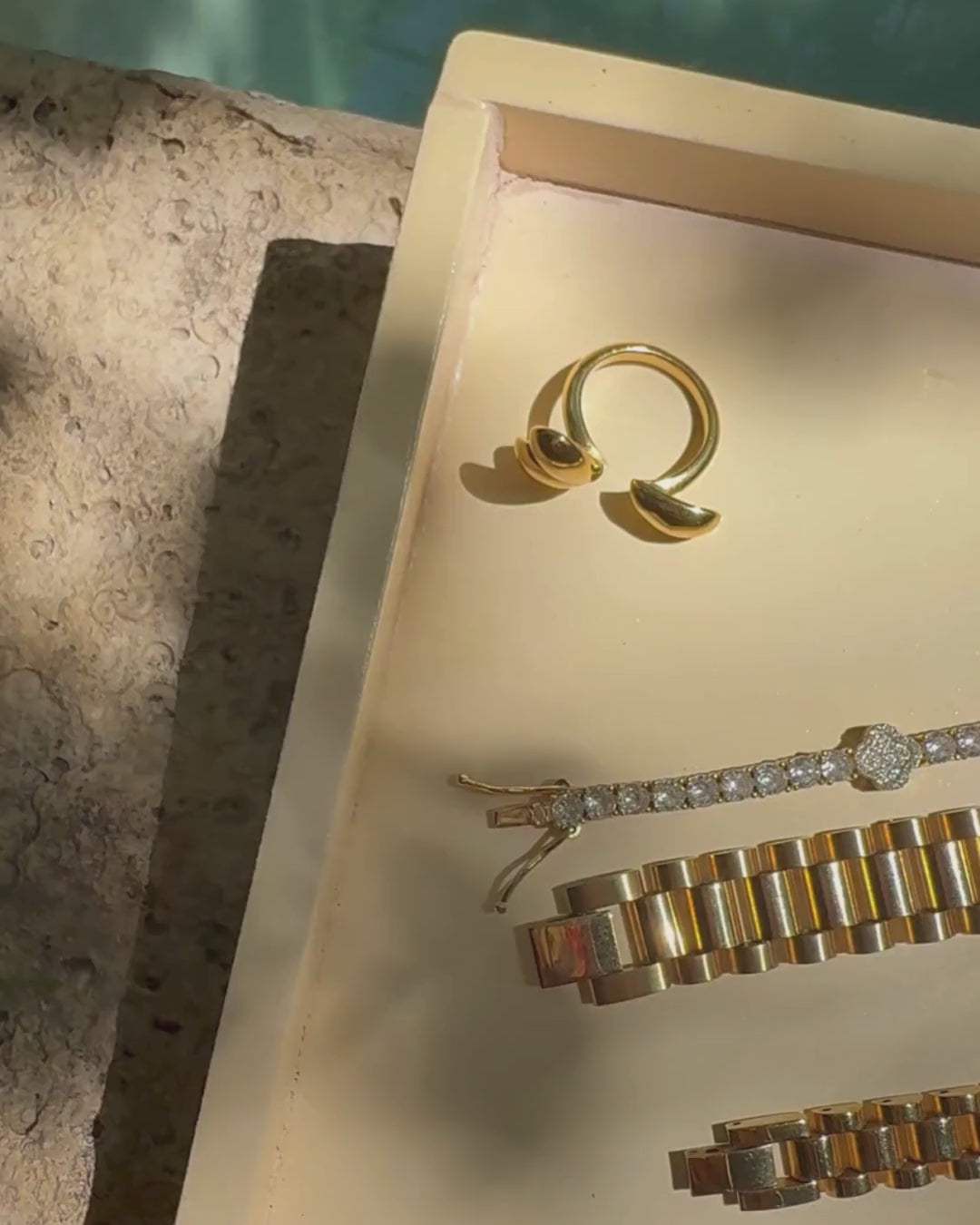 Video pan across a jewelry tray displaying the Modern Muse Ring in an open tear-drop design, styled alongside SneakyLink Rolex-style bracelets and the wave-shaped ring, Wave-Runner Ring, for a bold luxury accessories showcase.